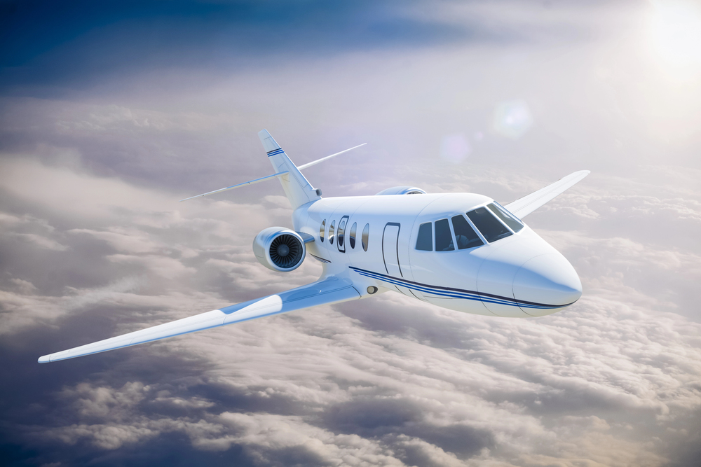 Small airplane parked on a concrete apron near a beige hangar and office building, with a fuel truck and cars in the background at a rural airfield.