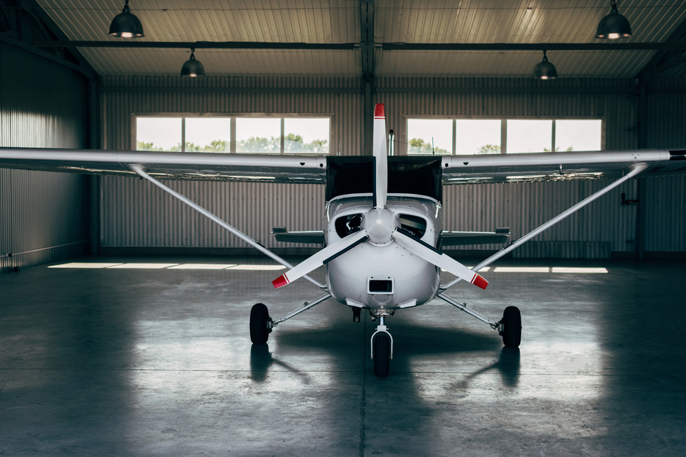 Small airplane parked on a concrete apron near a beige hangar and office building, with a fuel truck and cars in the background at a rural airfield.