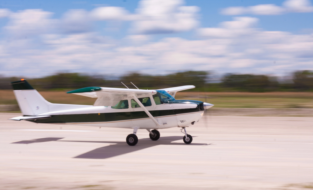 Small airplane parked on a concrete apron near a beige hangar and office building, with a fuel truck and cars in the background at a rural airfield.
