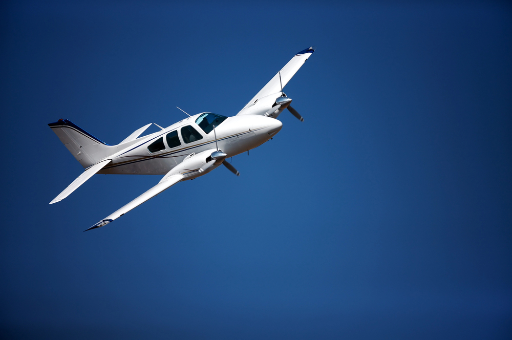 Small airplane parked on a concrete apron near a beige hangar and office building, with a fuel truck and cars in the background at a rural airfield.