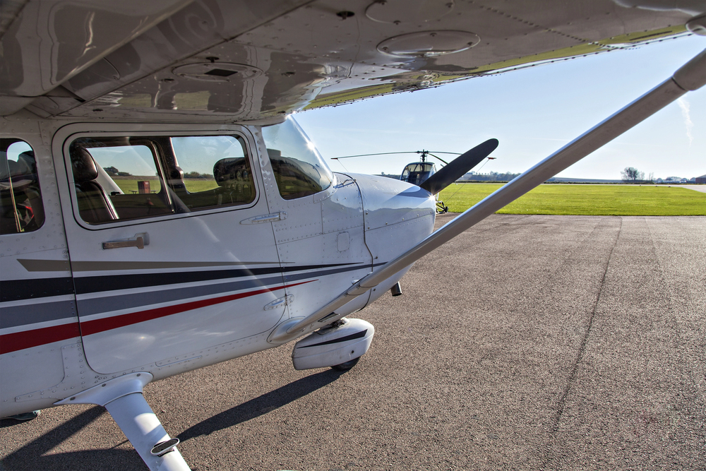 Small airplane parked on a concrete apron near a beige hangar and office building, with a fuel truck and cars in the background at a rural airfield.